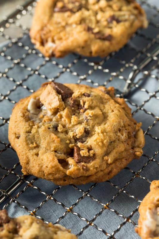 A close-up of a freshly baked smores cookie with chocolate chunks and a golden-brown texture, resting on a metal cooling rack over a blue cloth. Crumbs and soft edges enhance its homemade appearance, inviting you to try this delightful cookie recipe.