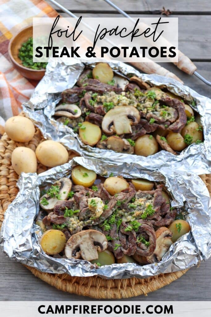 Foil packets filled with slices of steak, baby potatoes, mushrooms, and garnished with herbs. The packets are placed on a round woven mat with a cloth nearby and a small bowl of chopped herbs in the background. Text reads Foil Packet Steak & Potatoes.