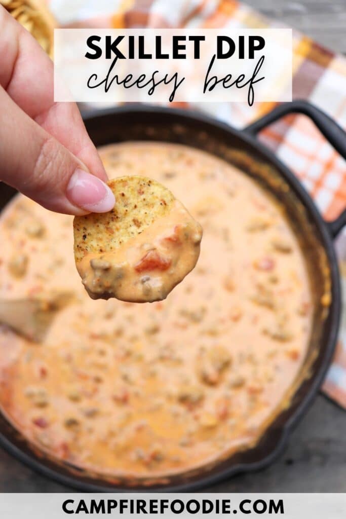 A hand holding a chip dipped in a creamy, beefy cheese sauce over a skillet filled with more of the dip. A checkered cloth is partially visible in the background. The text reads Skillet Dip Cheesy Beef and campfirefoodie.com.