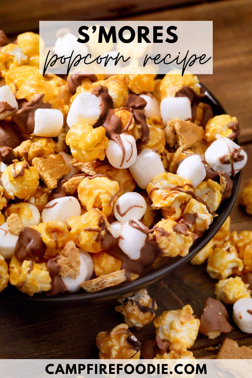 A black bowl overflowing with smores popcorn, featuring caramel popcorn, mini marshmallows, graham cracker pieces, and chocolate drizzle. Text overlay reads S’mores popcorn recipe and campfirefoodie.com at the bottom.