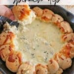 A skillet filled with creamy spinach dip surrounded by a ring of baked bread rolls. A hand is dipping a rol in the cheese. Topped with melted cheese, the dish is set on a wooden surface with a checkered cloth nearby.