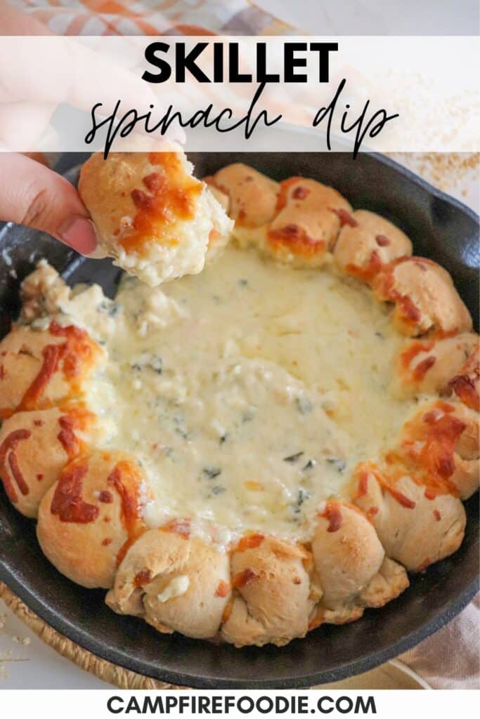 A skillet filled with creamy spinach dip surrounded by a ring of baked bread rolls. A hand is dipping a rol in the cheese. Topped with melted cheese, the dish is set on a wooden surface with a checkered cloth nearby.