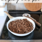 A frying pan on a stove contains cooked ground taco meat. A wooden spoon rests on the pans handle. The background shows a portion of a kitchen counter with a cutting board. Text at the top reads Taco Meat Camping Meal Plan.