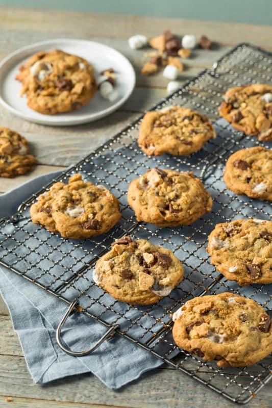 Freshly baked smores cookies with chocolate chips and nuts rest on a cooling rack atop a wooden table draped with a blue cloth. A plate with a couple of these delightful cookies and scattered chocolate pieces hints at the tasty recipe in the background.