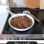 A white skillet filled with cooked taco meat sits on a stovetop. A wooden spoon rests on the skillets handle. In the background, there are cutting boards and a potted plant. Text at the bottom reads taco meat, CAMPING MEALS, campfirefoodie.com.