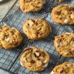 A cooling rack holds several freshly baked smores cookies loaded with chocolate chunks and marshmallows. They rest on a blue cloth-covered wire rack on a wooden surface, evoking the perfect smores cookie recipe. A partial view of a plate with more cookies is in the background.