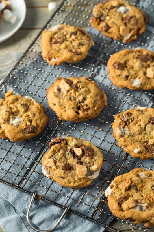 A cooling rack holds several freshly baked smores cookies loaded with chocolate chunks and marshmallows. They rest on a blue cloth-covered wire rack on a wooden surface, evoking the perfect smores cookie recipe. A partial view of a plate with more cookies is in the background.