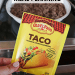 A hand holds an Old El Paso taco seasoning packet in front of a pan with cooking ground beef on a stove. The text reads ground beef meal planning and campfirefoodie.com.