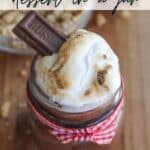 A dessert jar with layers of chocolate and toasted marshmallow, topped with a piece of chocolate. The jar is tied with a red and white checkered ribbon. Text overlay reads, Smores dessert in a jar.