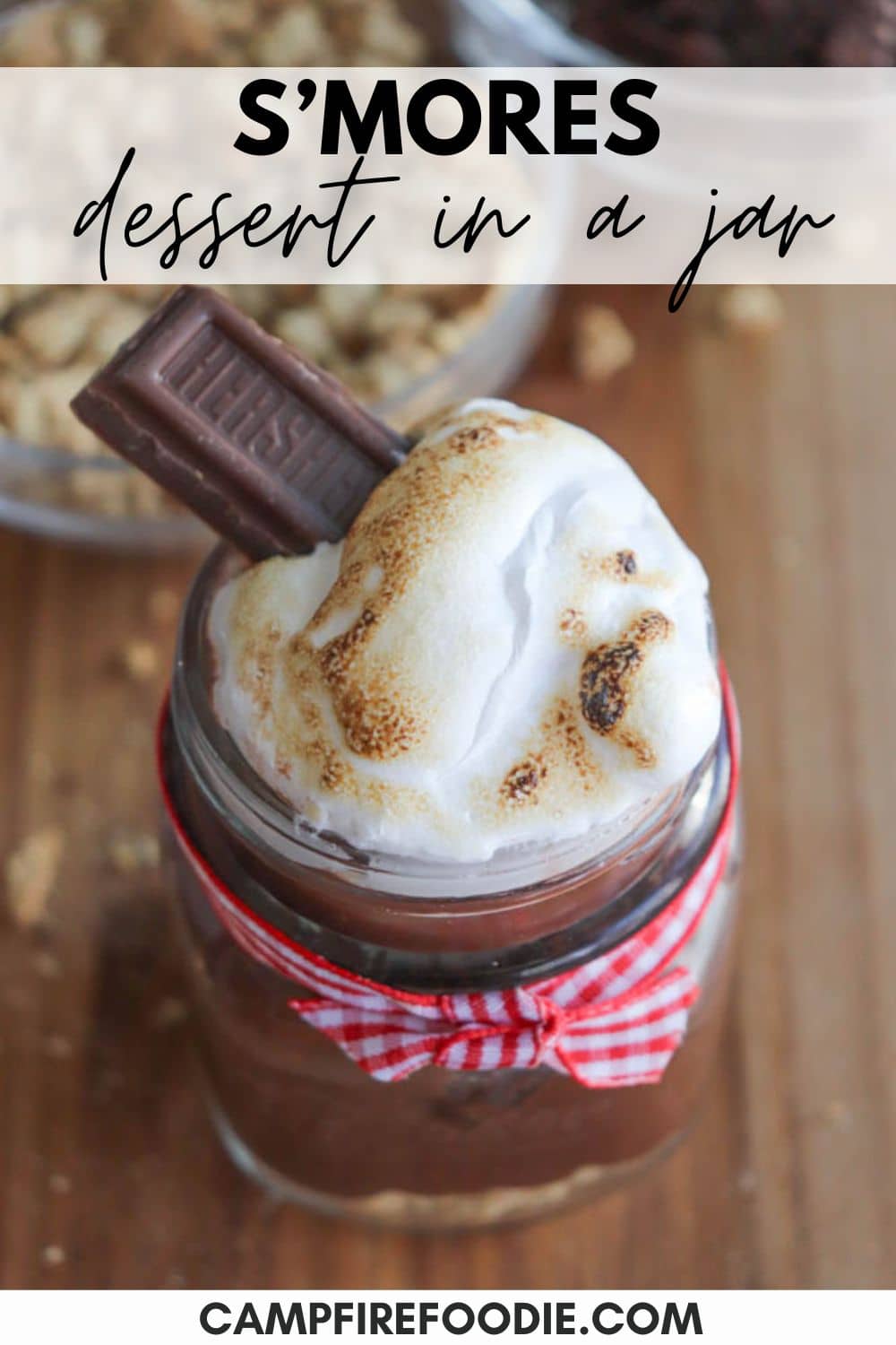 A dessert jar with layers of chocolate and toasted marshmallow, topped with a piece of chocolate. The jar is tied with a red and white checkered ribbon. Text overlay reads, Smores dessert in a jar.