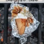 A waffle cone filled with melted marshmallows and chocolate is wrapped in foil and resting on a grill over a campfire. The text reads SMORES campfire cone at the top and CAMPFIREDOODIE.COM at the bottom.