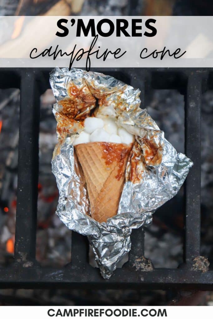 A waffle cone filled with melted marshmallows and chocolate is wrapped in foil and resting on a grill over a campfire. The text reads SMORES campfire cone at the top and CAMPFIREDOODIE.COM at the bottom.