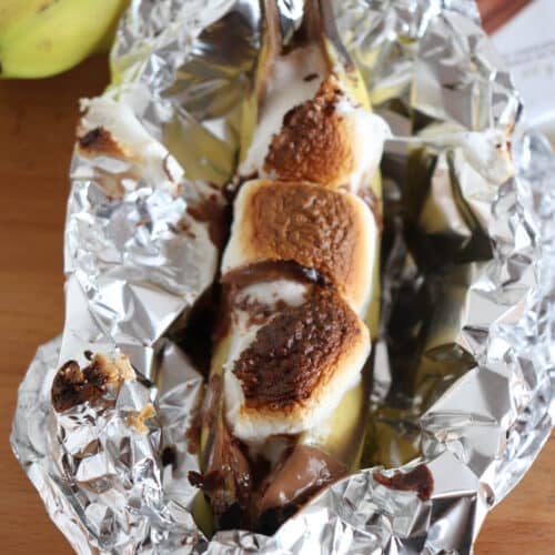 A banana split open and topped with melted chocolate and toasted marshmallows, wrapped in aluminum foil. There are whole bananas and a chocolate bar in the background on a wooden surface.