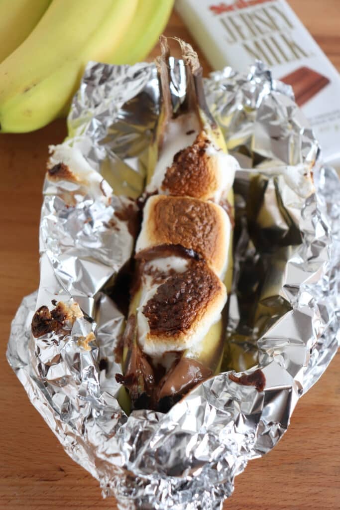 A banana split open and topped with melted chocolate and toasted marshmallows, wrapped in aluminum foil. There are whole bananas and a chocolate bar in the background on a wooden surface.