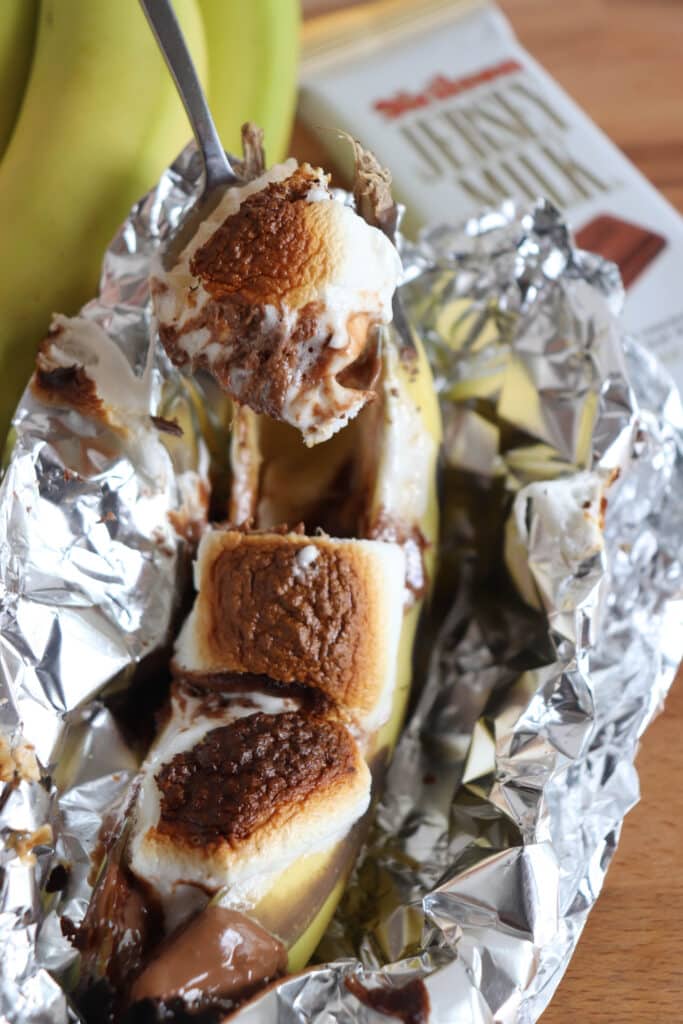 A foil-wrapped banana with melted chocolate and toasted marshmallows, held over with a spoon. A chocolate wrapper and bananas are in the background on a wooden table.