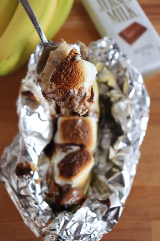 A spoon holds a bite of banana topped with melted chocolate and toasted marshmallows. The banana, chocolate, and marshmallows are arranged in foil, with a bar of chocolate and bananas visible in the background.