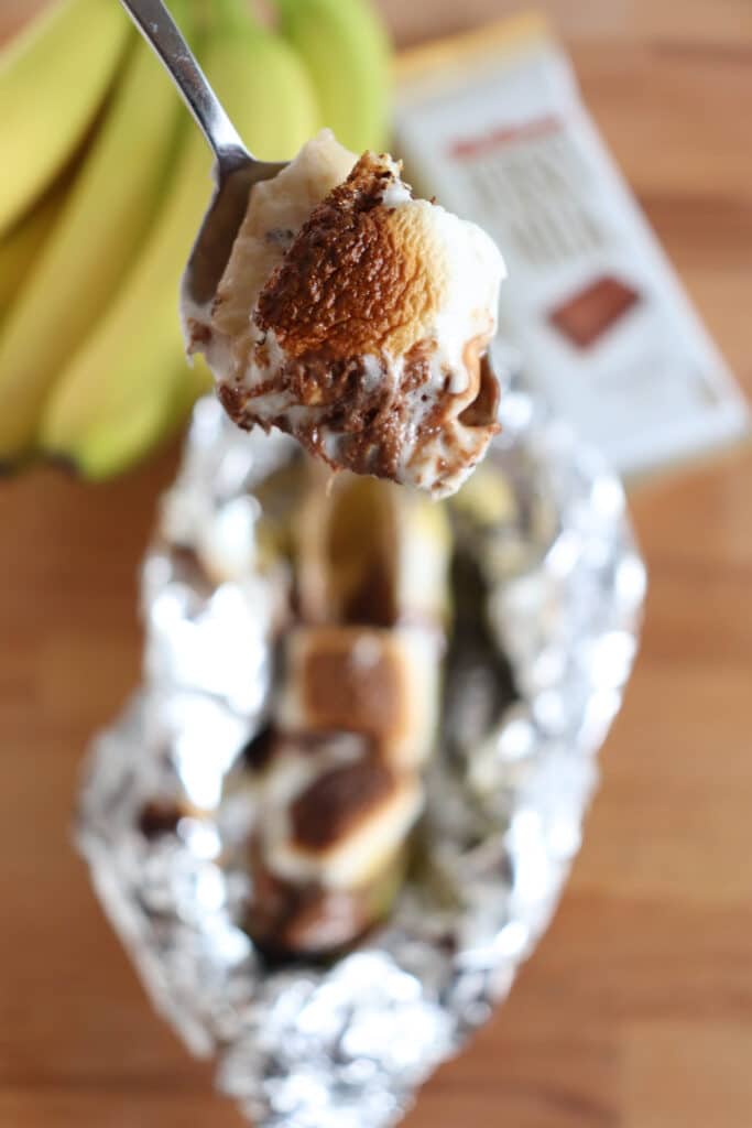 A spoon holds a bite of roasted marshmallow and chocolate over a foil-wrapped smores banana boat. In the background, theres a bunch of bananas and a partially visible chocolate bar on a wooden surface.