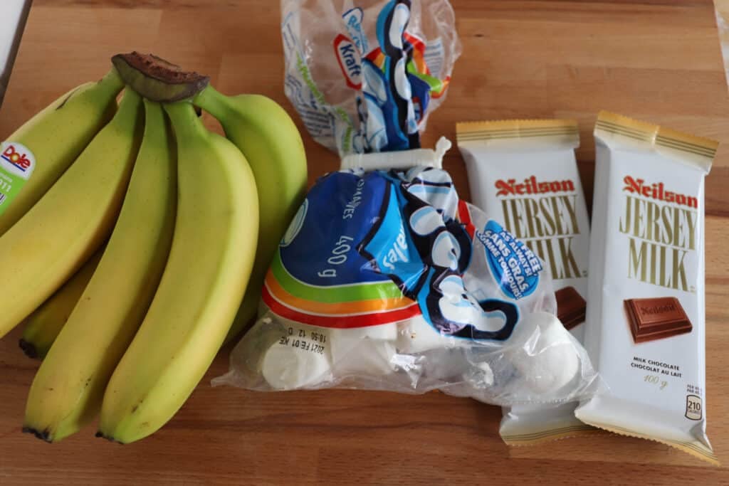 A bunch of bananas, a bag of marshmallows, and two bars of Neilson Jersey Milk chocolate are on a wooden surface.