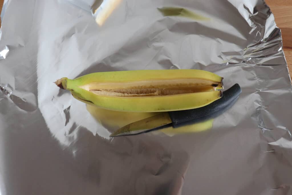 A partially peeled banana with a slit cut down the middle rests on silver foil. A small knife is placed beside the banana on the foil.