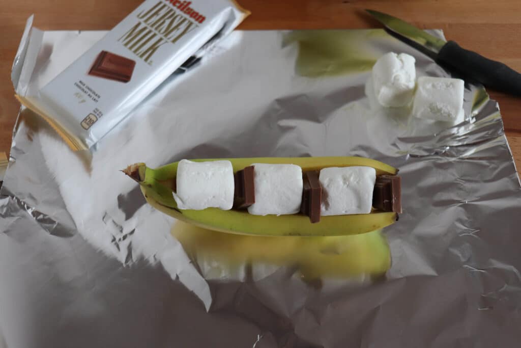 A banana sliced open lengthwise sits on aluminum foil, filled with pieces of milk chocolate and large marshmallows. A knife and an opened chocolate bar wrapper are visible in the background.