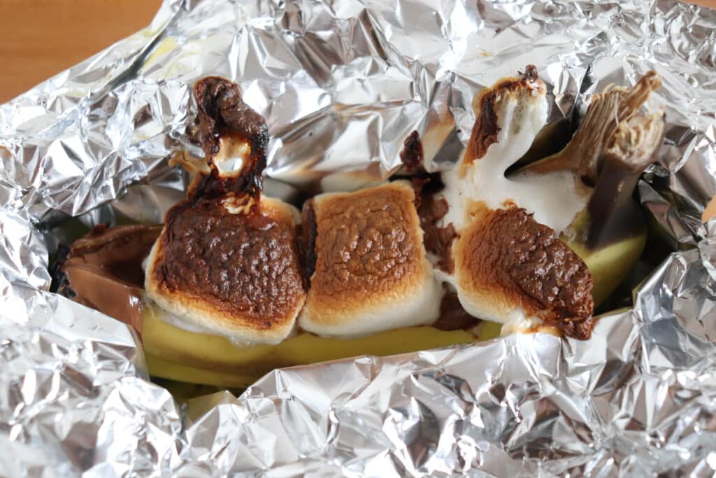 A banana split open and filled with roasted marshmallows and melted chocolate sits wrapped in crinkled aluminum foil. The marshmallows are toasted to a golden brown, and the chocolate is slightly melted.