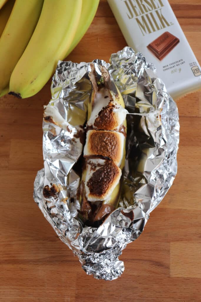 A peeled banana is topped with melted chocolate and toasted marshmallows, sitting on aluminum foil. Nearby are a bunch of bananas and a bar of Jersey Milk chocolate on a wooden surface.