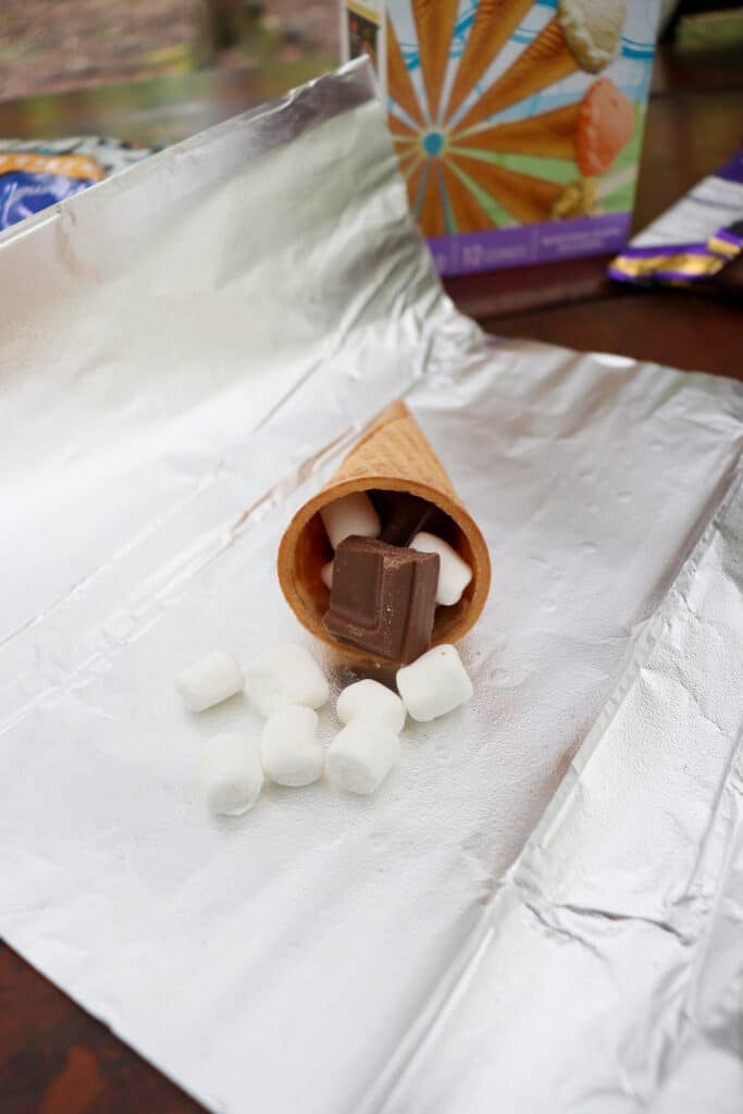 A waffle cone filled with chocolate and marshmallows is placed on a piece of aluminum foil. Additional mini marshmallows are scattered outside the cone. A colorful box is slightly visible in the background.