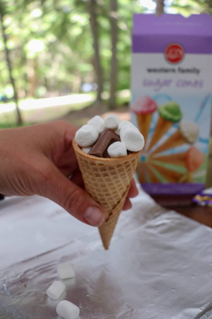 A hand holds a waffle cone filled with mini marshmallows and a chocolate piece. In the background, a box with images of colorful ice cream cones is partially visible. The scene is set outdoors with blurred green trees.