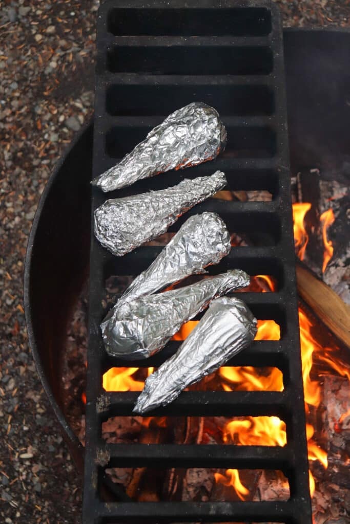 Four food items wrapped in aluminum foil are placed on a grill over a campfire. The flames and embers are visible beneath the grill, and the surrounding area is scattered with small stones and gravel.