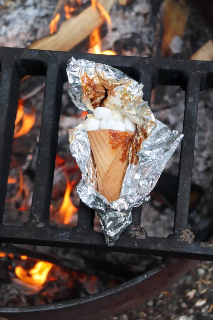 A waffle cone wrapped in foil is filled with marshmallows and placed on a grill over a campfire, with flames visible below.