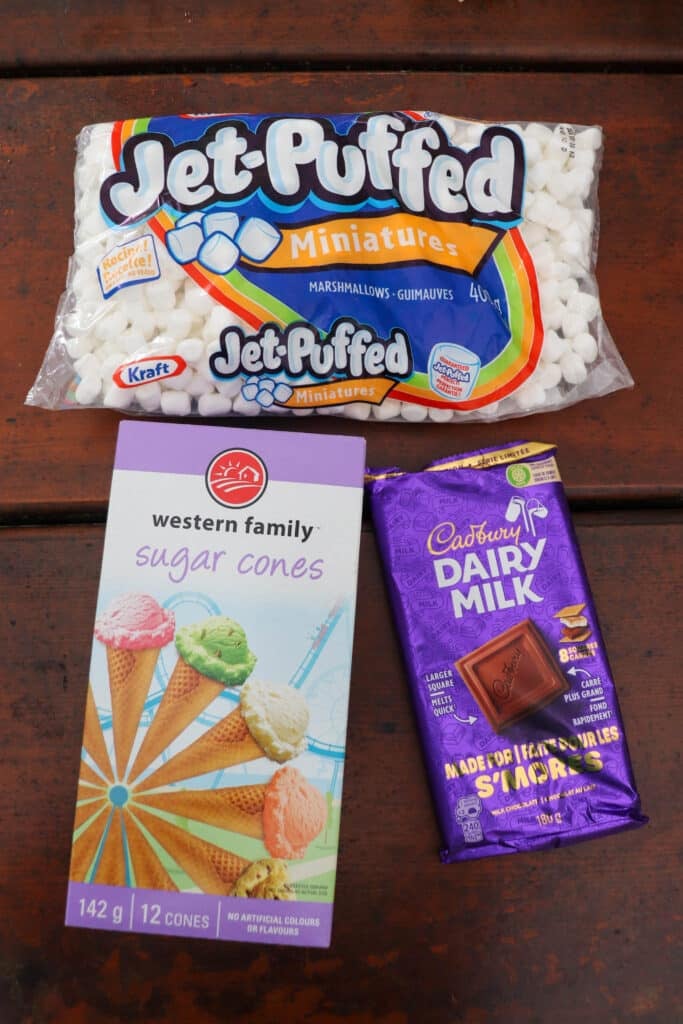 A package of Jet-Puffed Miniature Marshmallows, a box of Western Family Sugar Cones featuring colorful ice cream images, and a Cadbury Dairy Milk chocolate bar are laid out on a wooden surface.