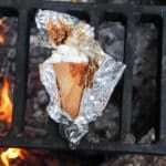 A waffle cone filled with marshmallows and chocolate, wrapped in aluminum foil, sits on a grill over an open flame. The foil is partially opened, revealing some melted marshmallows and chocolate.