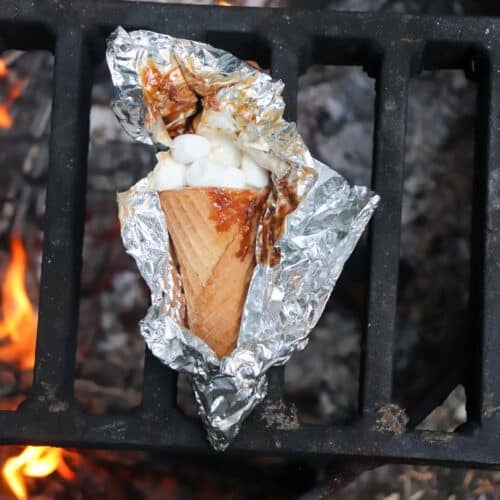 A waffle cone filled with marshmallows and chocolate, wrapped in aluminum foil, sits on a grill over an open flame. The foil is partially opened, revealing some melted marshmallows and chocolate.