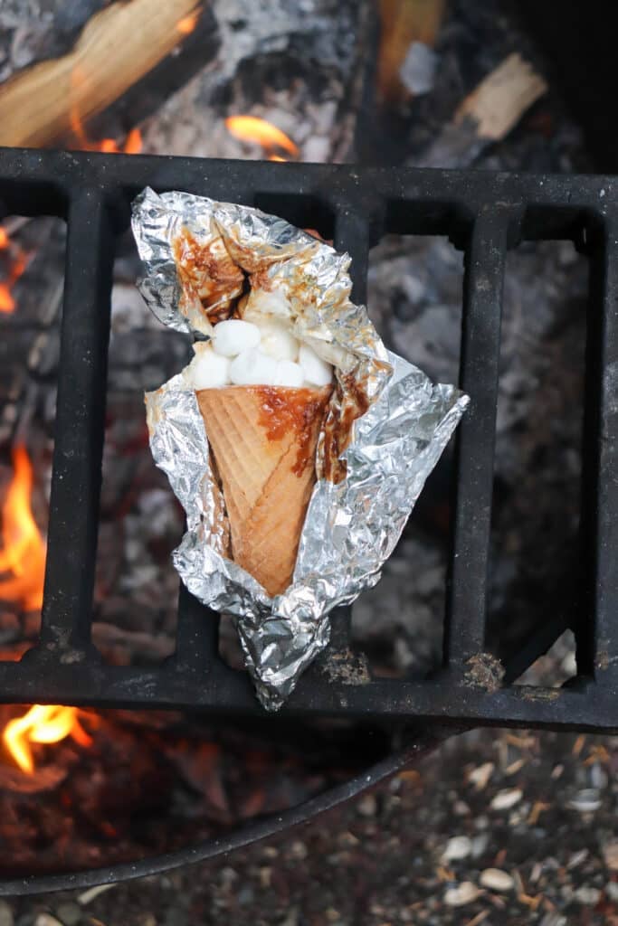 A waffle cone filled with marshmallows and chocolate, wrapped in aluminum foil, sits on a grill over an open flame. The foil is partially opened, revealing some melted marshmallows and chocolate.