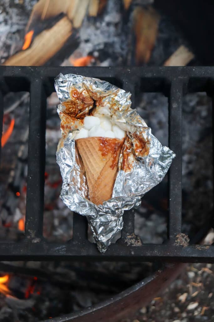 A waffle cone filled with marshmallows and chocolate is partially wrapped in aluminum foil, sitting on a grill over an open flame. The background shows glowing embers and wood pieces.