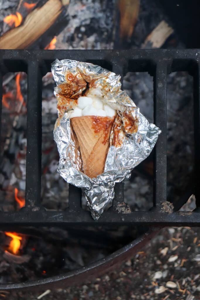 A waffle cone filled with marshmallows, wrapped in aluminum foil, is placed on a grill above a fire. The flames and charred wood create a warm, outdoor cooking scene.