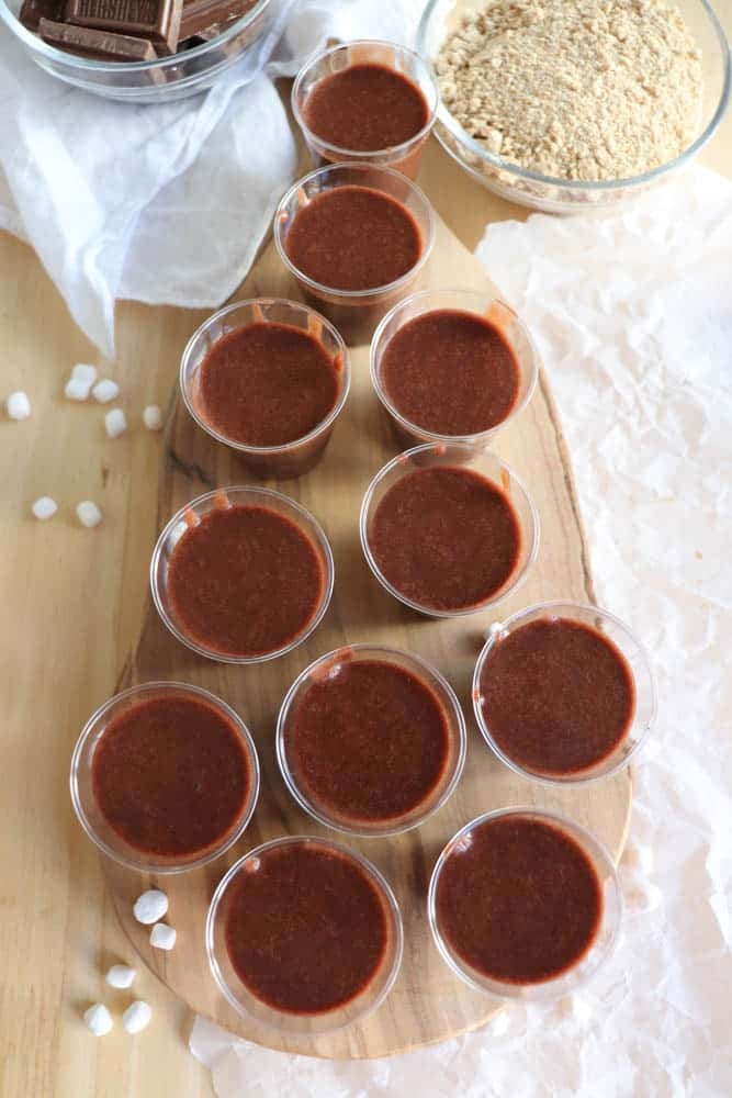 Overhead view of ten plastic cups filled with chocolate liquid on a wooden board. Surrounding items include a bowl of crushed graham crackers, chocolate pieces, and small marshmallows scattered on a light wooden table.