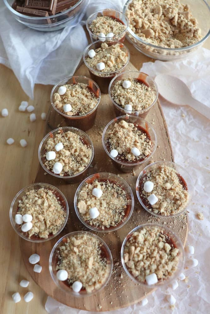 A wooden board holds nine small cups of dessert, each topped with crumbled cookies and mini marshmallows. A bowl of crumbled cookies and a bar of chocolate are visible in the background. The setup is on a light wooden surface.