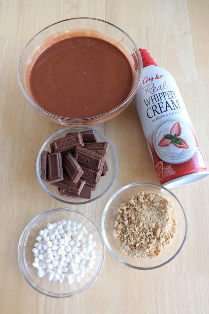 Bowls of chocolate pudding, chocolate pieces, mini marshmallows, and crushed cookies are on a table, along with a can of whipped cream.