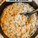 A black pot filled with creamy macaroni and cheese, featuring spiraled pasta and melted cheese. A serving spoon rests in the dish. The text at the top reads Dutch Oven Mac & Cheese and at the bottom campfirefoodie.com.