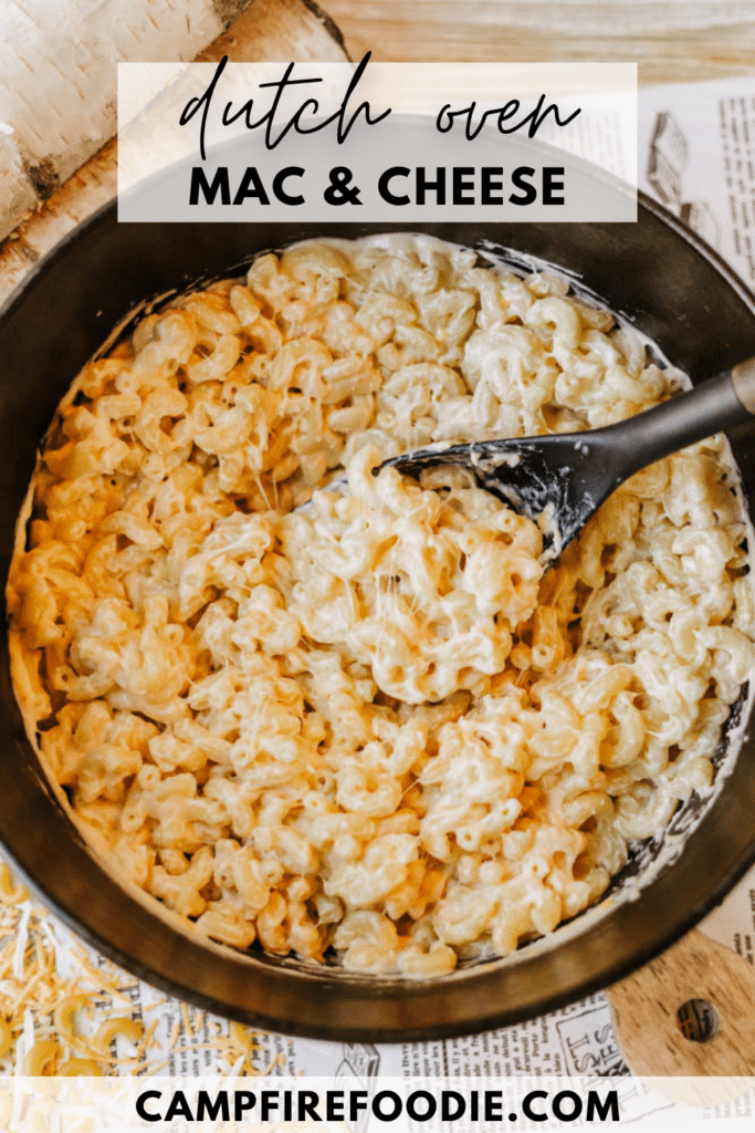 A black pot filled with creamy macaroni and cheese, featuring spiraled pasta and melted cheese. A serving spoon rests in the dish. The text at the top reads Dutch Oven Mac & Cheese and at the bottom campfirefoodie.com.