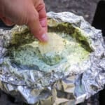 A hand holds a tortilla chip, dipping it into creamy campfire spinach dip nestled in a foil packet. The packet sits on the grill, and the dip looks hot and freshly prepared.