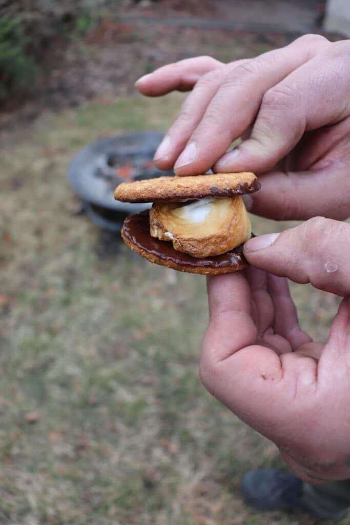 Hands holding the best smores, with perfectly toasted marshmallow and rich chocolate sandwiched between two crunchy graham crackers, set against a lush grassy background.