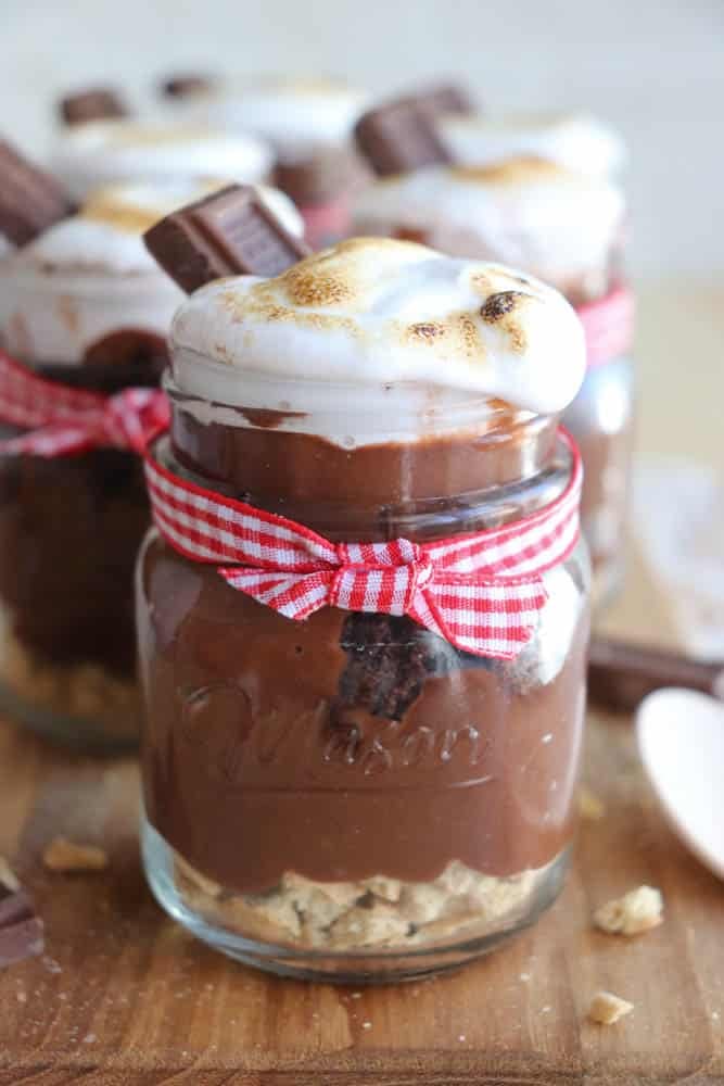 A dessert jar filled with graham cracker crumbs, chocolate pudding, and topped with toasted marshmallow fluff. The jar is tied with a red-and-white checkered ribbon, and a piece of chocolate is inserted into the topping. Other jars are blurred in the background.