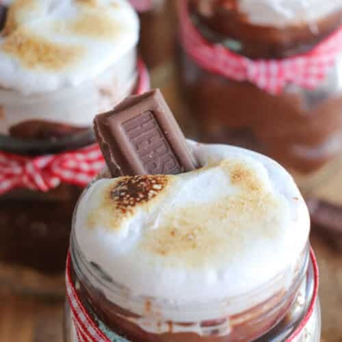 Close-up of mason jars filled with chocolate dessert, topped with toasted marshmallows and a piece of chocolate. The jars are decorated with red and white striped ribbon.