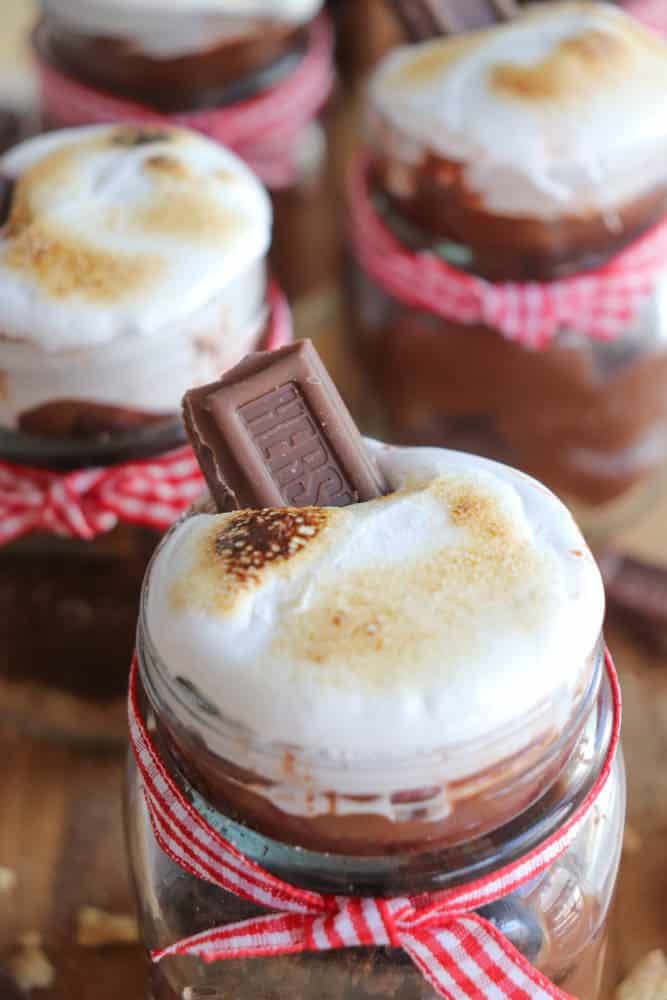 Close-up of mason jars filled with chocolate dessert, topped with toasted marshmallows and a piece of chocolate. The jars are decorated with red and white striped ribbon.