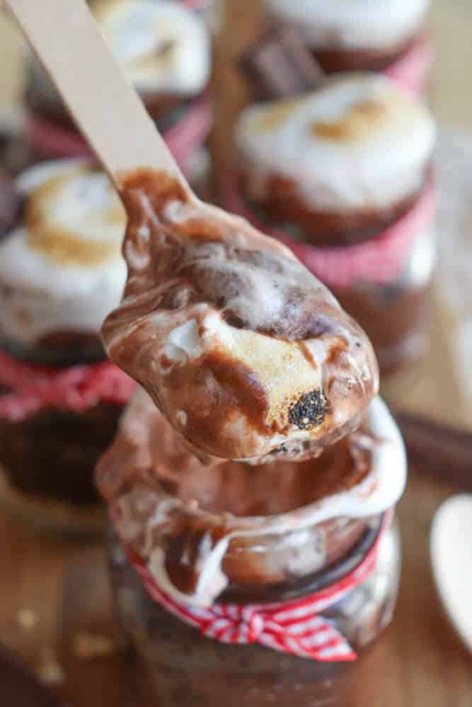 A wooden spoon holds up a serving of chocolate dessert with toasted marshmallow on top. Below, glass jars filled with more of the dessert are decorated with red and white ribbons.