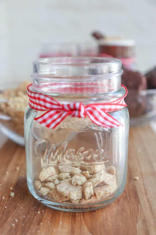 A glass mason jar with a red and white checkered ribbon around the neck, containing small cereal pieces. The jar is on a wooden surface, with blurred jars and snacks in the background.