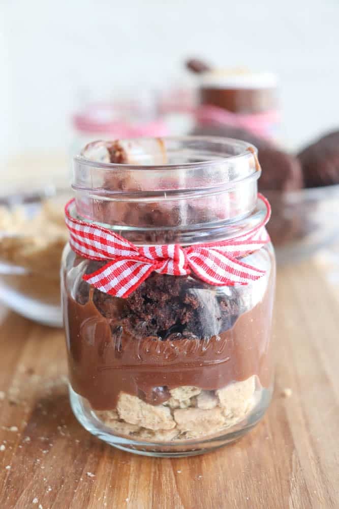 A glass jar filled with layered dessert: crushed cookies at the bottom, followed by chocolate pudding and brownie pieces on top. The jar is tied with a red and white checkered ribbon. It sits on a wooden surface, with a similar jar blurred in the background.