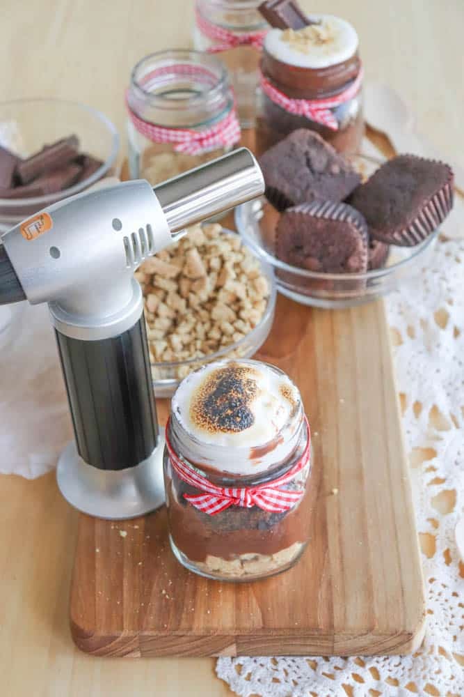 A spread of dessert ingredients on a wooden board, featuring a blowtorch, jars with layered desserts topped with toasted marshmallow, chopped nuts, chocolate, and small chocolate muffins. A lace cloth is partially visible beside the board.